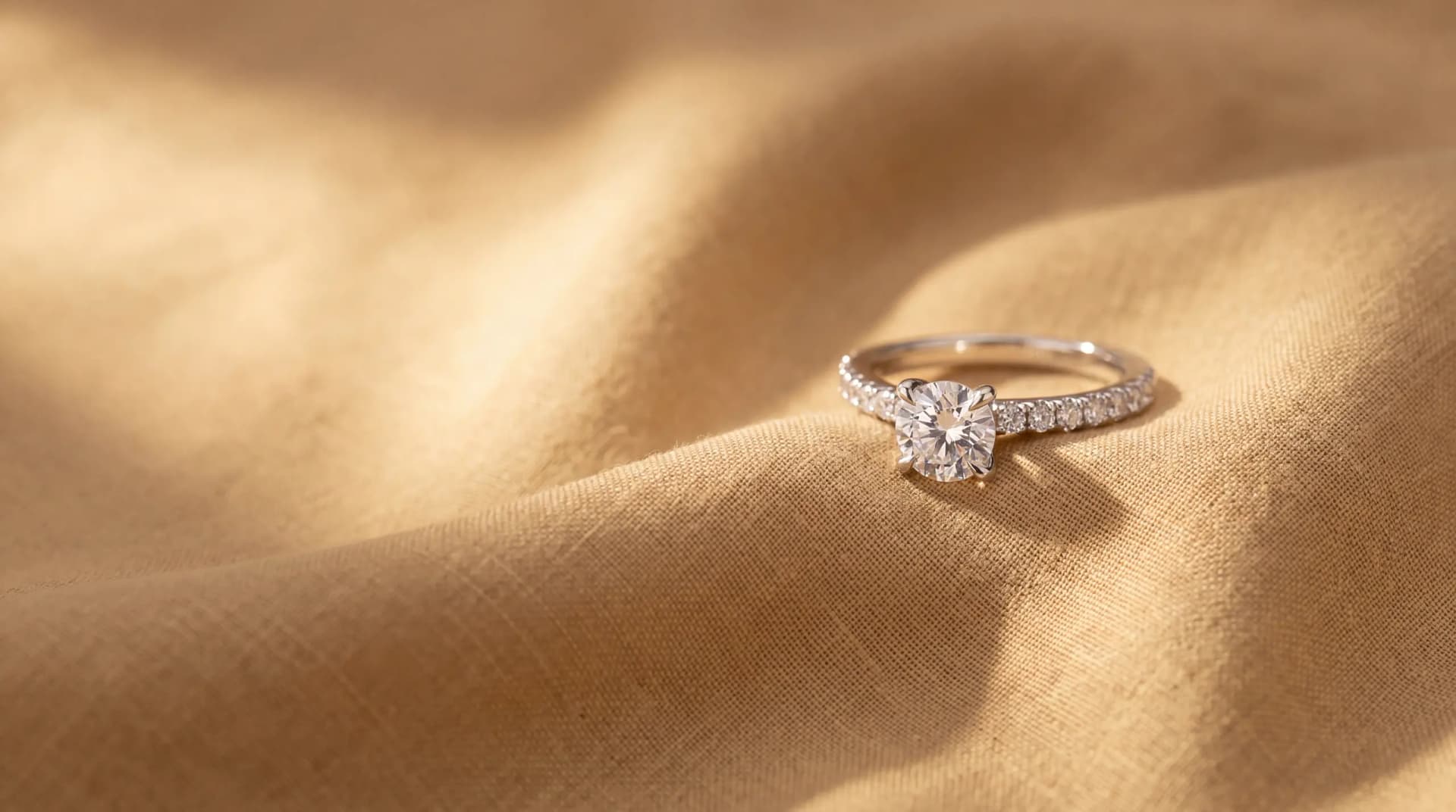 White gold solitaire engagement ring, its round brilliant centre diamond held in four claws above a shank set with small round diamonds, resting on champagne linen