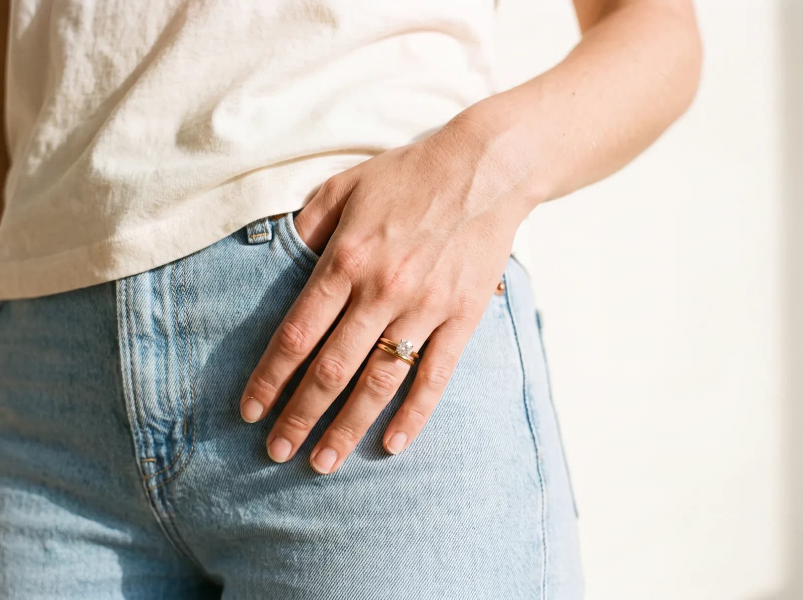 A hand tucked into a denim jean pocket, wearing a round solitaire ring and a plain stacking band, a cream t-shirt visible above.