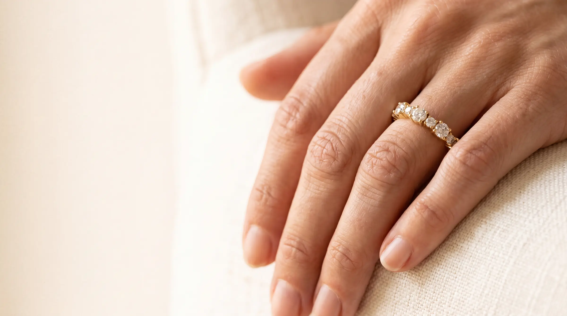 A woman's hand resting on top of her other hand on sunlit ivory fabric, wearing a yellow gold ring set with a row of round diamonds across the band, with the left two-thirds of the frame empty sunlit fabric.